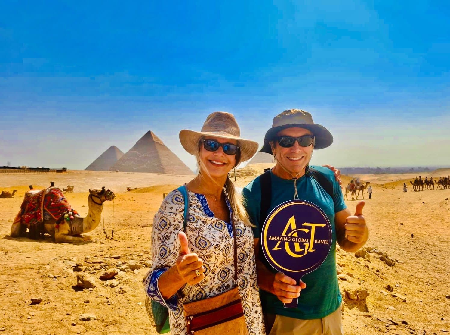 Two travellers holding the Amazing Global Travel sign beside a camel, the Giza pyramids behind them